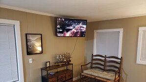 A living room with a television mounted on the wall, a wooden bench, a small wooden cabinet, and a patterned rug. - Camp Fishbone - Our place at the Lake (Freeport)