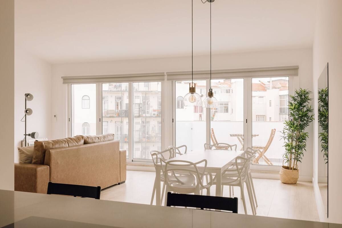 A bright living room with a sofa, dining table, chairs, and large windows offering a view of buildings.