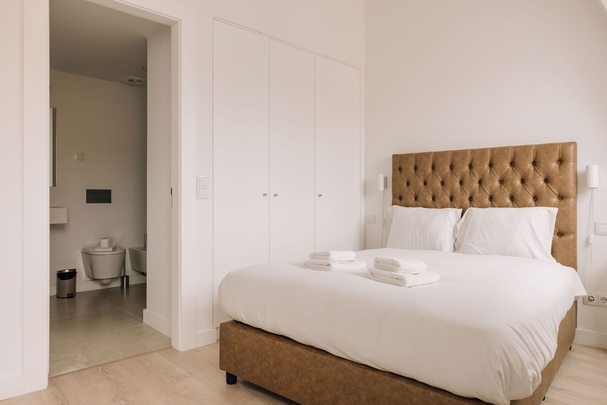A bedroom with a large bed, white bedding, a tufted headboard, and a view of the bathroom through an open door.