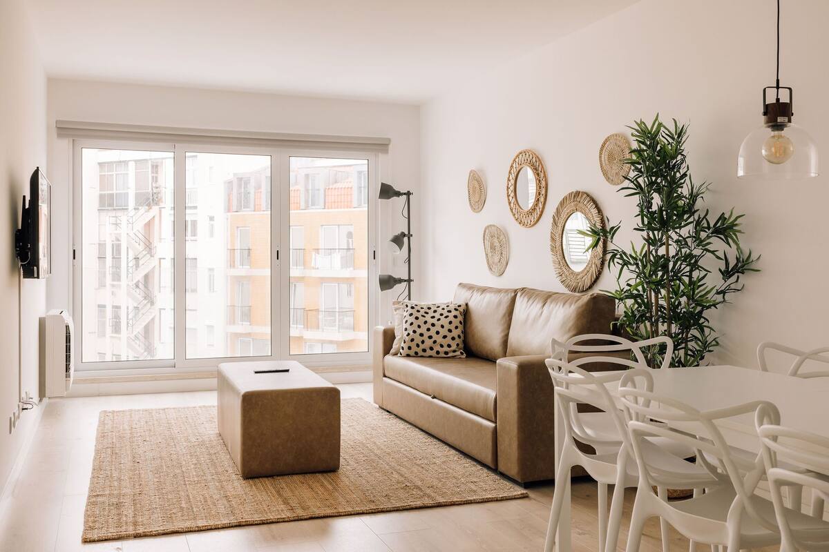A modern living room with a sofa, ottoman, dining table, chairs, and a large window.