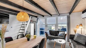 A modern living room with a wooden dining table, a sofa, and a coffee table. There is a staircase leading to an upper level, and large windows providing natural light. - Cozy apartment in Hemmet with sauna (Hemmet)