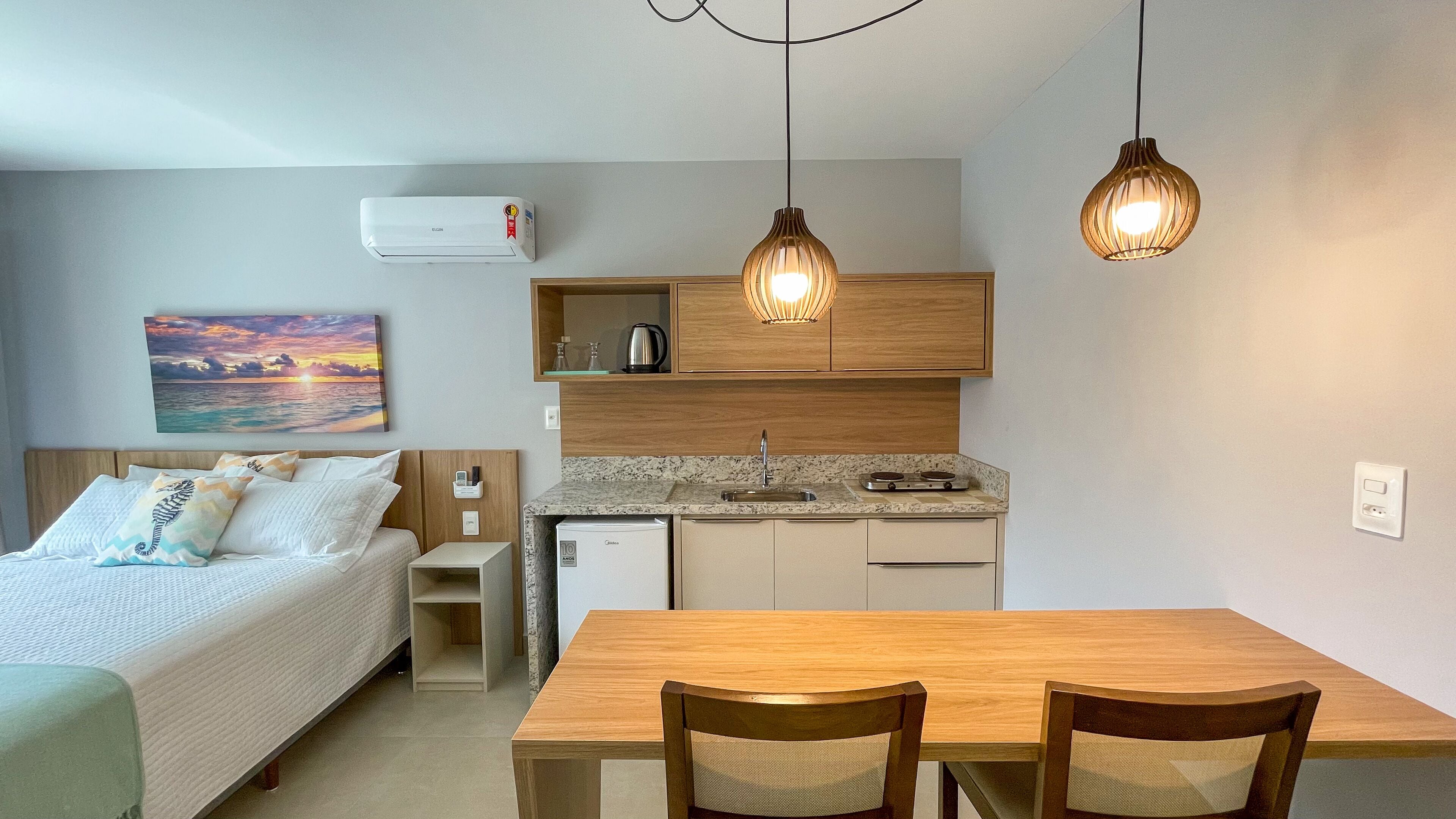 A compact hotel room with a bed, bedside table, a small kitchenette, a dining area with a table and chairs, and a painting of a sunset on the wall.