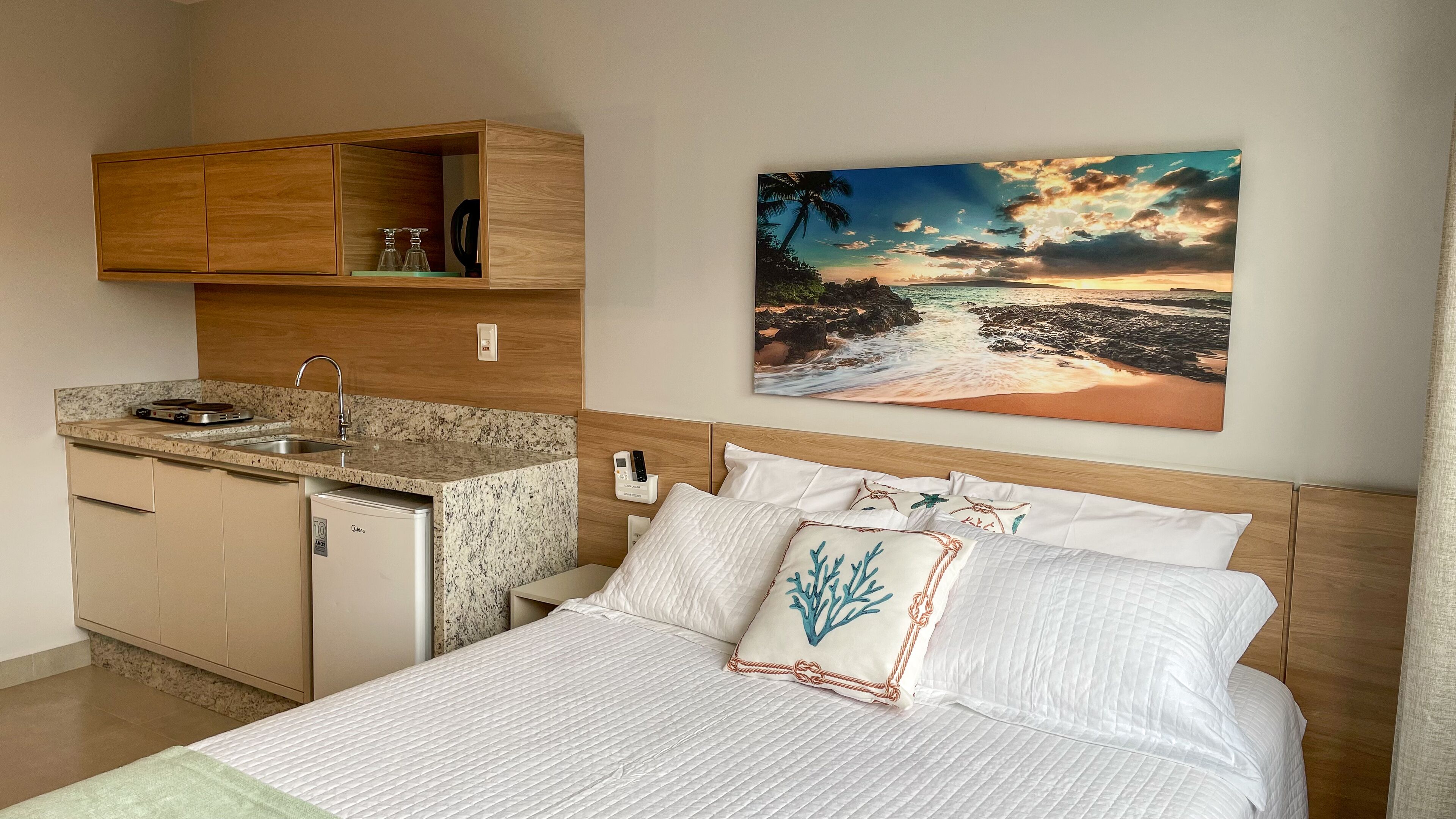 A hotel room with a bed, a small kitchenette, a painting of a beach scene, and a wooden headboard.