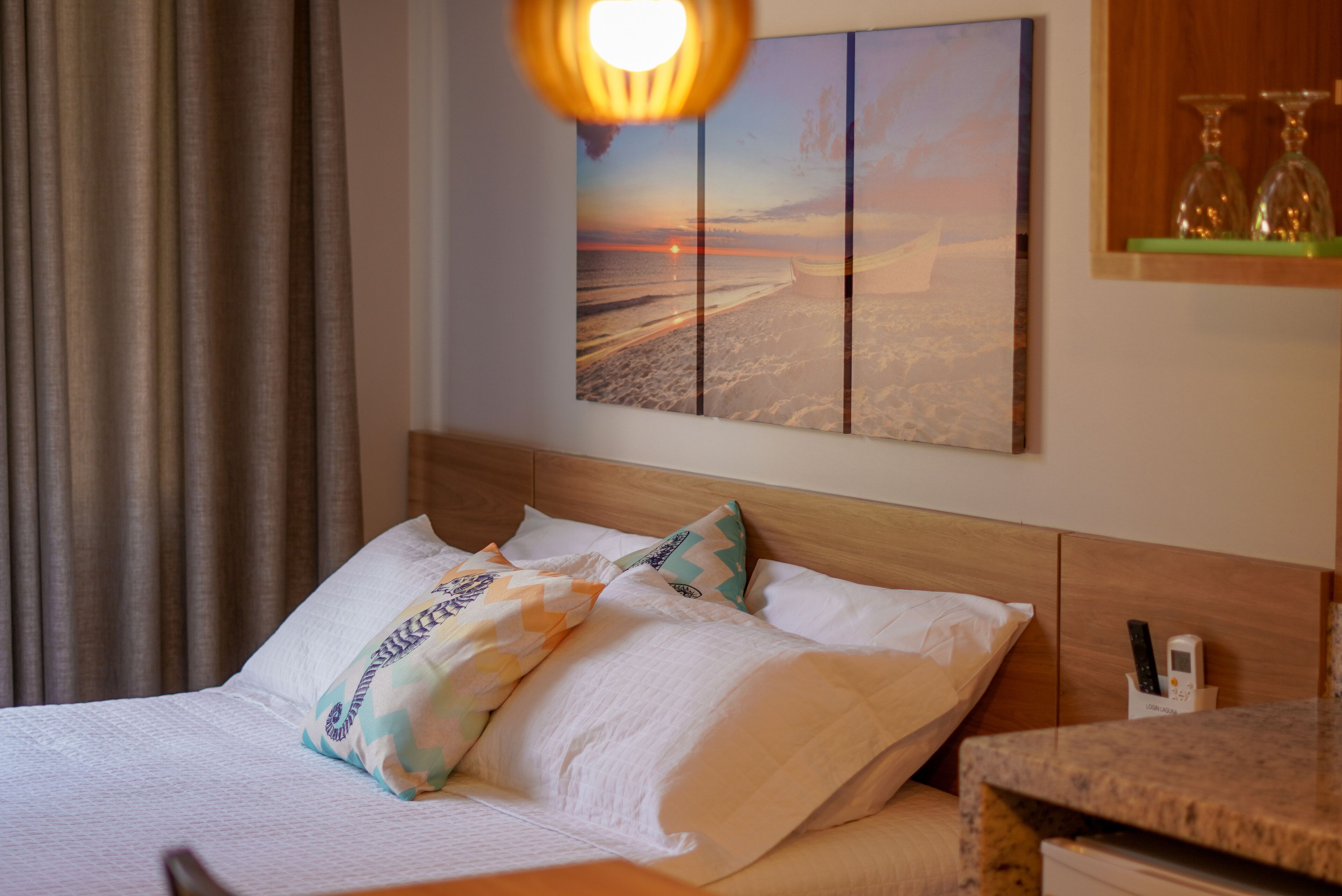 A neatly made bed with patterned pillows, a wall-mounted painting of a beach scene, and a bedside table with a remote control.