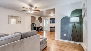 A modern living room with a grey sofa, wooden flooring, a ceiling fan, and a large mirror. - 15% Summer Discount~ Entire Home ~ Full Kitchen ~ Large Fenced Backyard (Oklahoma City)