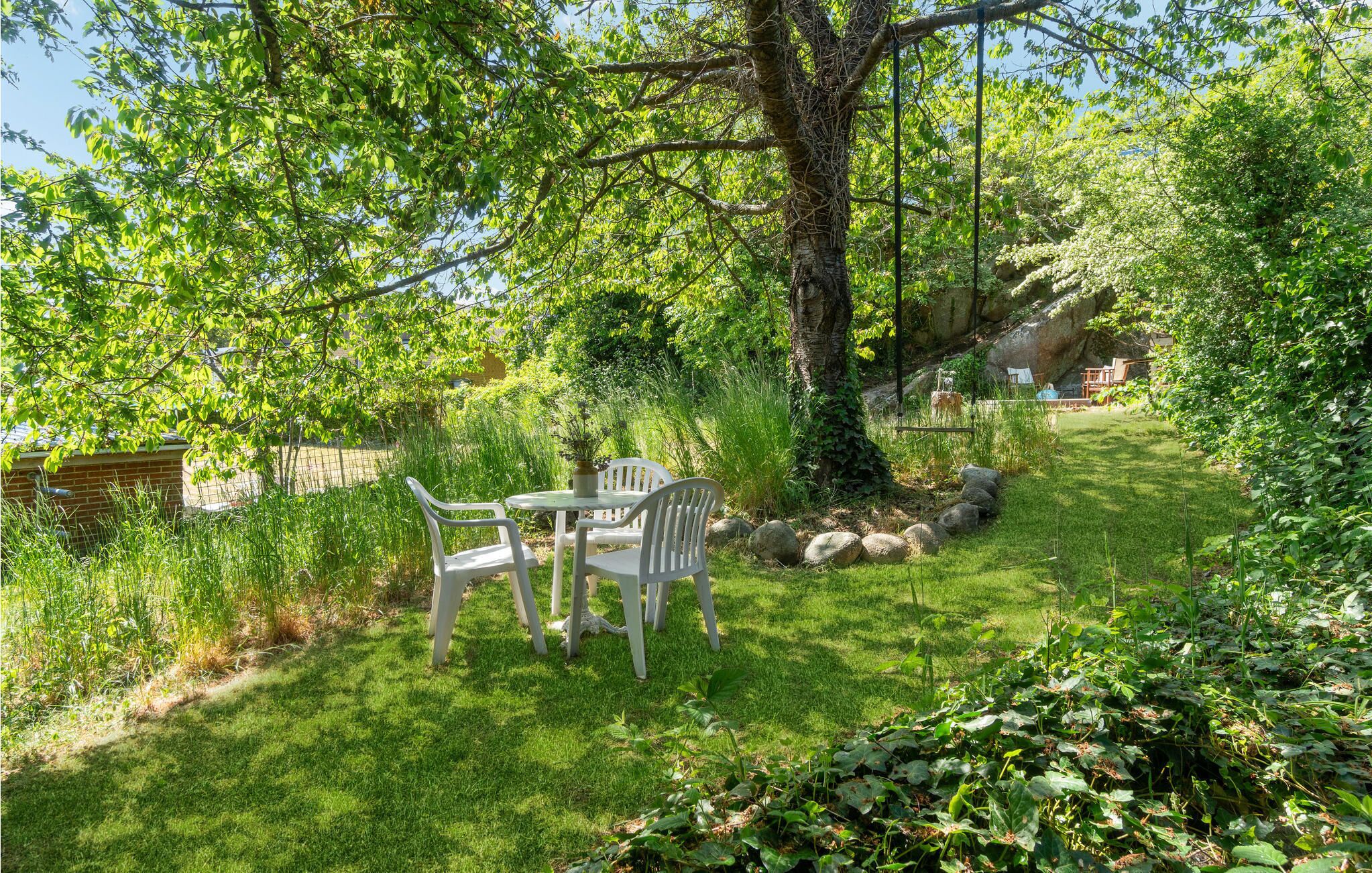 Un jardin avec une table et des chaises, entouré d’une végétation luxuriante et d’un grand arbre.