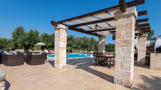 Un patio al aire libre con piscina, pilares de piedra, mobiliario de mimbre y una pérgola.
