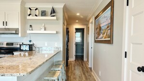 A kitchen with a granite countertop, white cabinets, and a wooden floor. There are bar stools and a refrigerator in the background. - Pet Friendly, Ocean Front w/ Heated Private Pool and Sun Deck (Brunswick)