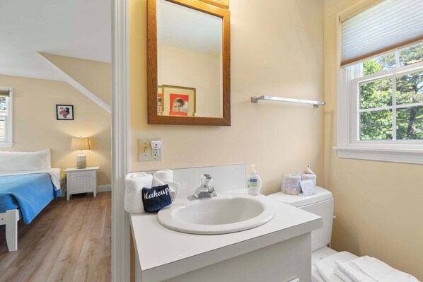 Une salle de bain avec un lavabo blanc, un miroir et des toilettes. Il y a un porte-serviettes au-dessus du lavabo et une fenêtre donnant sur des arbres.