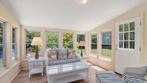 Una habitación soleada con un sofá, una mesa de centro y una silla. Hay ventanas con cortinas, una puerta y una lámpara sobre una mesita.