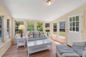 Ein sonnendurchflutetes Zimmer mit einer Couch, einem Couchtisch und einem Sessel. Es gibt Fenster mit Vorhängen, eine Tür und eine Lampe auf einem Beistelltisch.