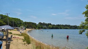Una playa junto al lago, con un paraguas a rayas, orilla arenosa y gente nadando.