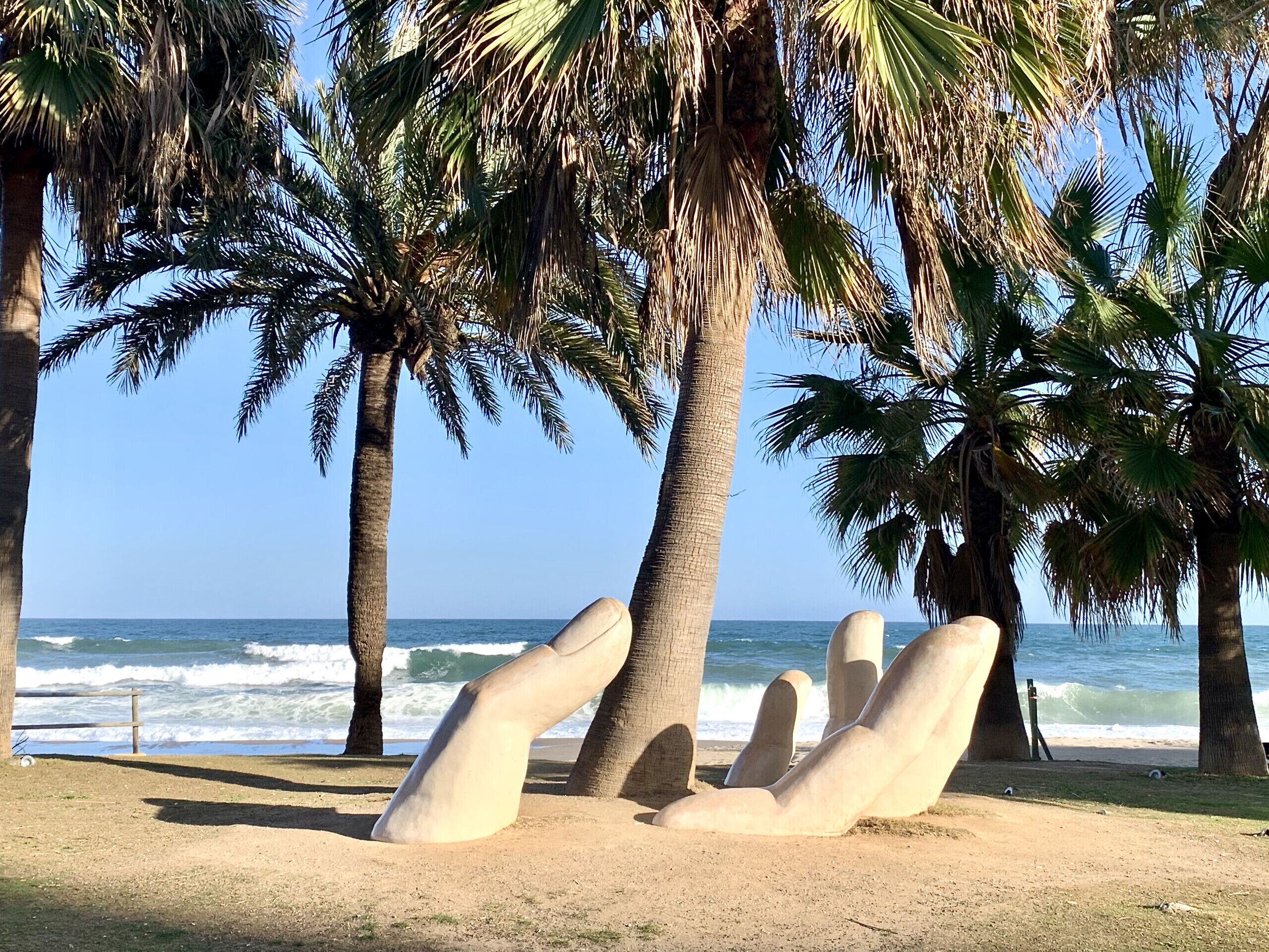 Une sculpture représentant des figures allongées dans un parc, avec des palmiers et une vue sur l’océan.