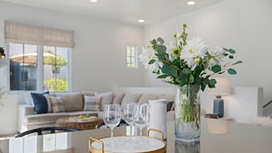 A modern living room with a glass coffee table, a vase with white flowers, and a white sofa with cushions. - Modern Luxury Condo In Uptown SB (Santa Barbara)