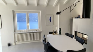 Een eethoek met een grote witte tafel, zwarte stoelen, een raam met uitzicht op zee, en een radiator eronder.
