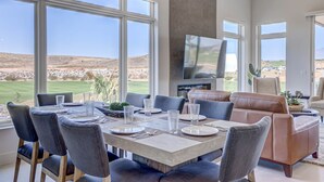 Un comedor con una mesa grande dispuesta para seis personas, rodeada de sillas, un televisor montado en la pared y vistas al exterior.