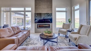 Amplia sala de estar con un sofá grande, una mesa de centro y dos sillones. Una chimenea está ubicada en el centro de la pared, y hay ventanas amplias que ofrecen vistas al exterior.