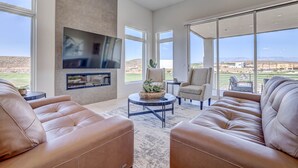 Amplia sala de estar con sofás de cuero, un televisor de pantalla plana montado en una pared texturizada y grandes ventanales con vistas a un campo de golf.