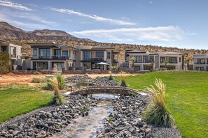 Un moderno edificio residencial de varios pisos con un jardín arbolado, un pequeño arroyo y vistas a montañas.