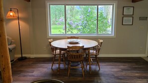 A dining area with a round table and chairs, a window with a view of greenery, and a floor lamp.