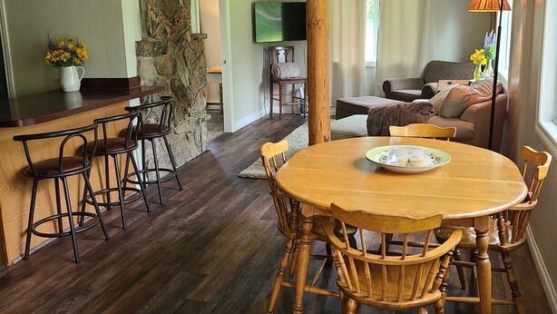 A dining area with a wooden table and chairs, a stone wall, a clock, and a television in the background.