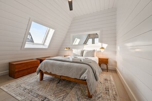 Quarto com teto inclinado, uma janela grande, cama com manta cinza, mesa de cabeceira de madeira e um criado-mudo com abajur.
