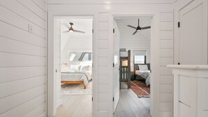 A hallway with white paneled walls and wooden flooring, leading to a bedroom with a bed and a ceiling fan.