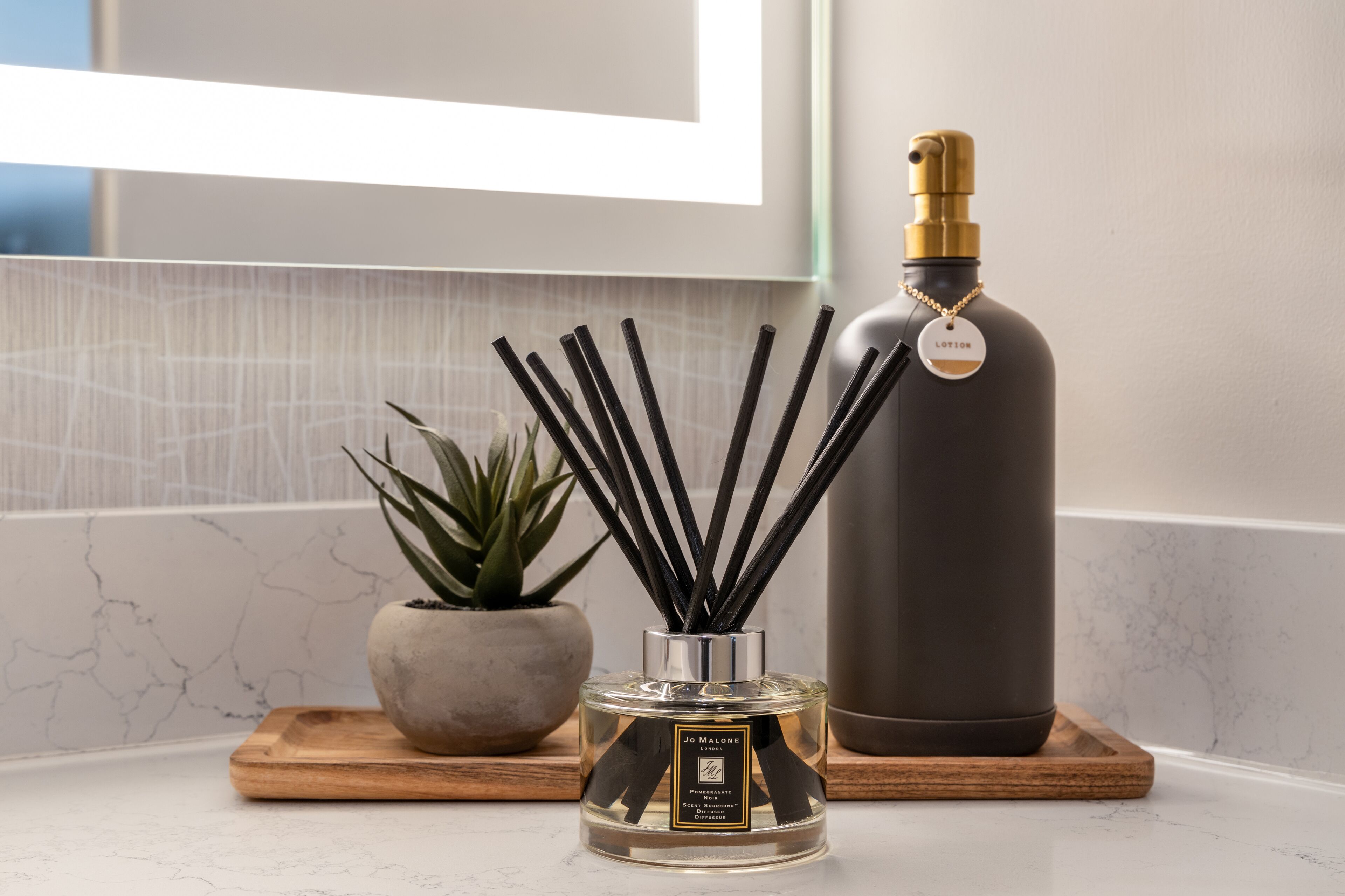 Um frasco de perfume sobre uma bandeja de madeira, um frasco de vidro com bastões pretos e uma planta em vaso.
