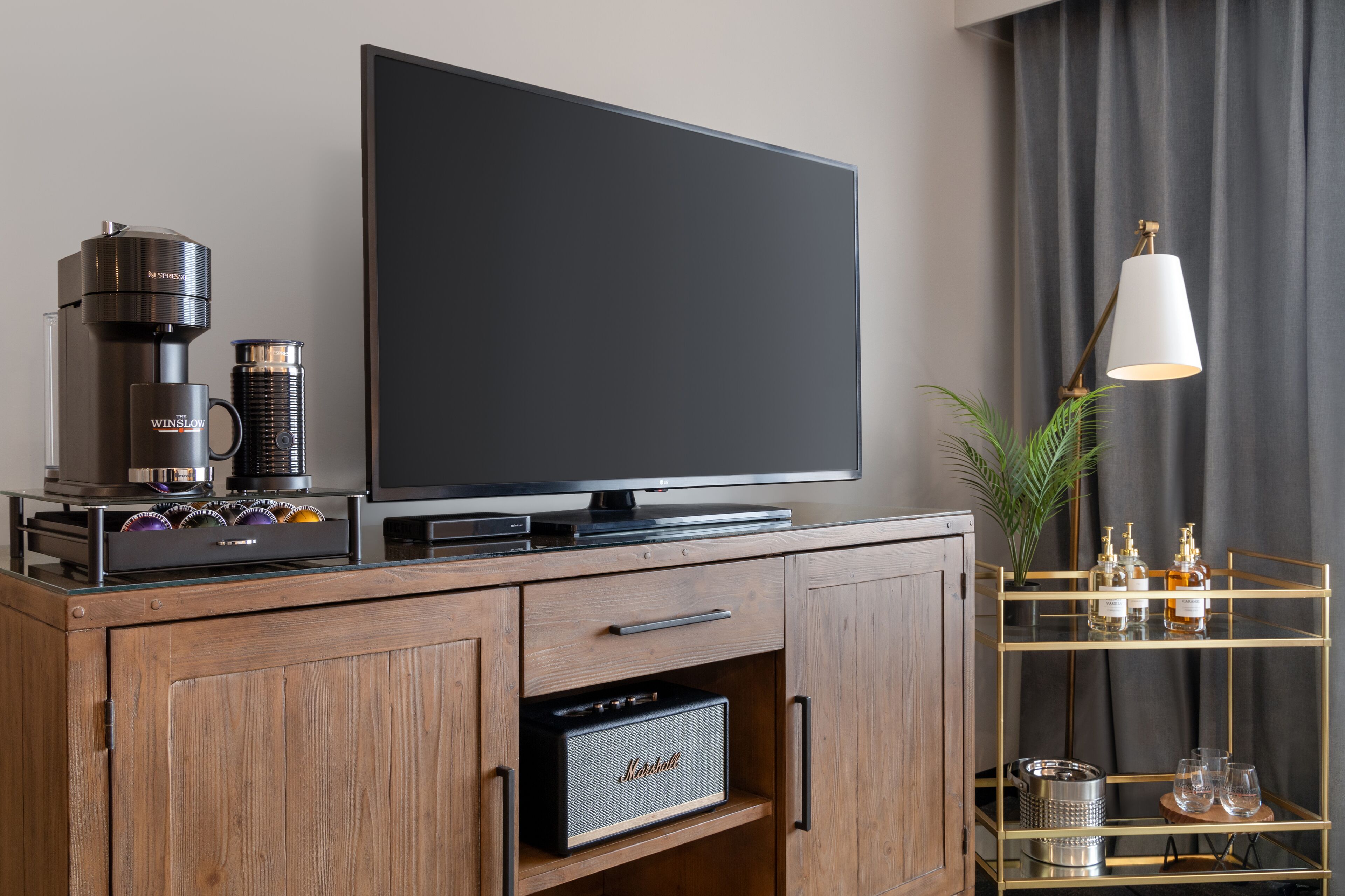 Una sala de estar moderna con un televisor de pantalla plana, una cafetera, un amplificador Marshall y una estantería con marco dorado que contiene botellas y vasos.