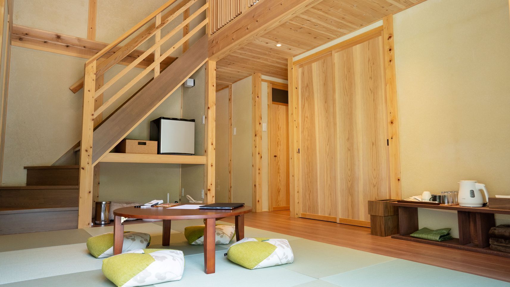 A room with a wooden staircase, a table with cushions, and a kettle on a small table.