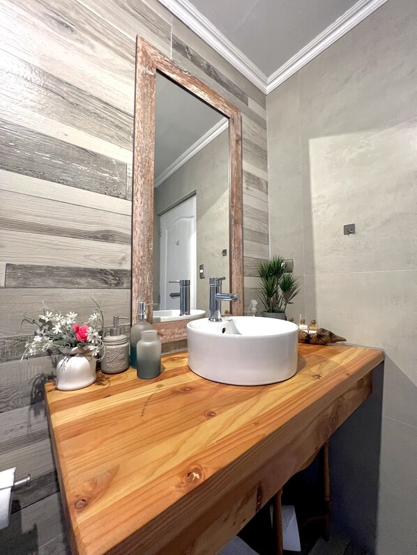 A bathroom with a wooden vanity, a white sink, a large mirror, and a potted plant. - Hare Mahatu (Hanga Roa)