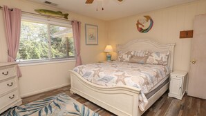 Ein Schlafzimmer mit Bett, Nachttisch, Kommode, einem Fenster mit Vorhängen, Deckenventilator und Wanddekoration.