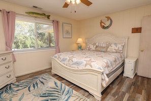 A bedroom with a bed, a nightstand, a dresser, a window with curtains, a ceiling fan, and a wall decoration.