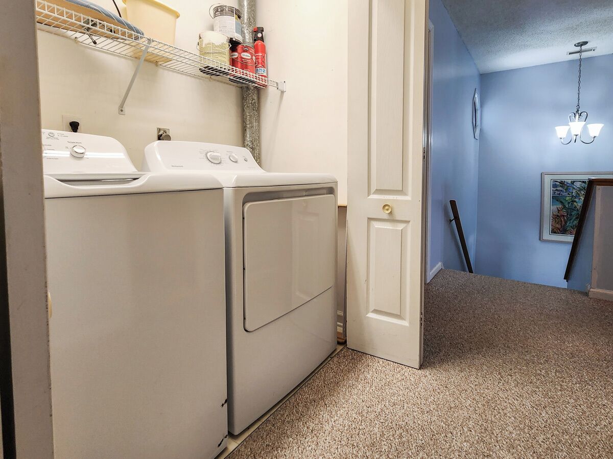 A laundry room with two white washers and dryers, a shelf with cleaning supplies, and a door leading to another room.