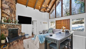 A cozy living room with a stone fireplace, wooden ceiling, and large windows.
