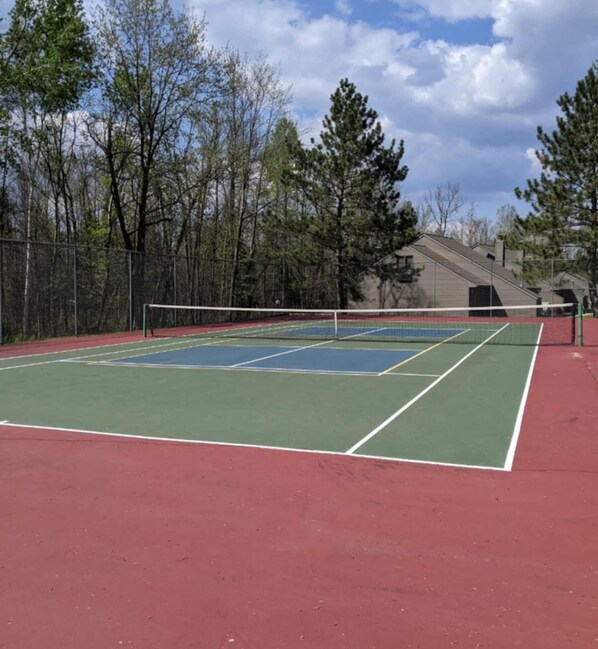 Una cancha de tenis al aire libre con una red, rodeada de árboles y un edificio al fondo.