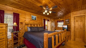 A wooden cabin bedroom with a bed, a ceiling fan, a TV, and a wooden door.