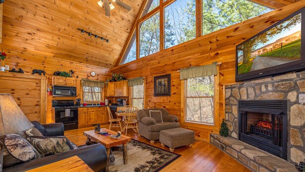 A cozy cabin interior with a stone fireplace, wooden furniture, and large windows.