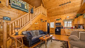 A log cabin interior with a wooden staircase, a sofa, a coffee table, a kitchen area, and a dining table.