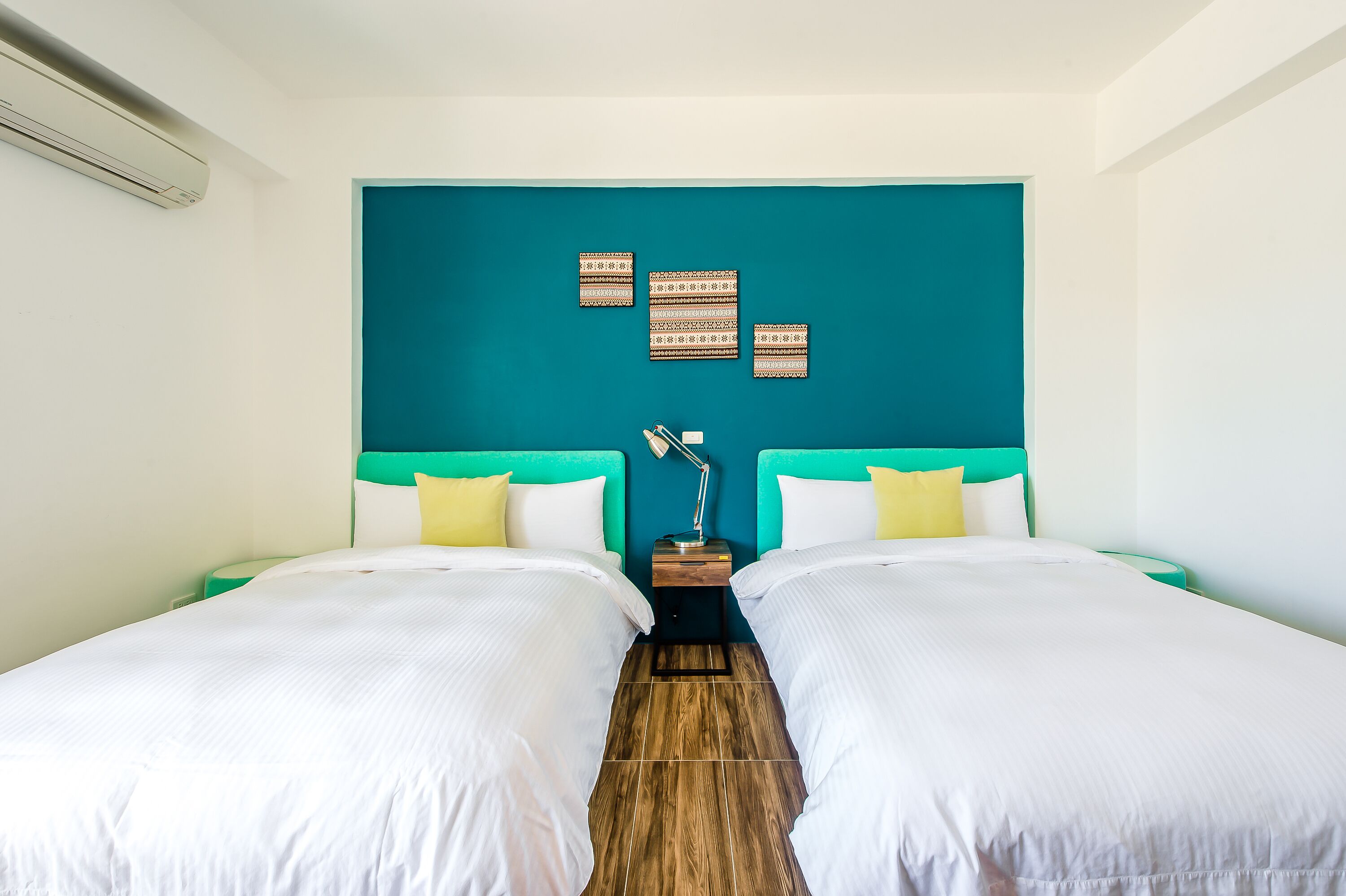 Habitación de hotel con dos camas, cabeceros color turquesa, ropa de cama blanca, una mesita de noche con lámpara y decoración en la pared.