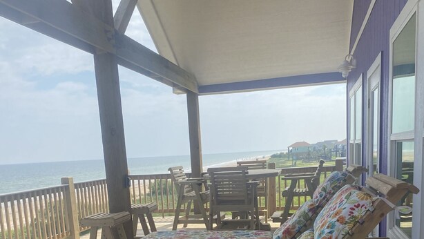 Una terraza de madera con una silla de descanso y una mesa, con vista a una playa y el océano.