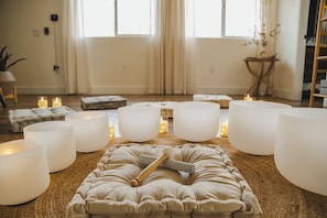 Una habitación con un otomano grande y mullido color beige en el centro, rodeado de almohadones blancos cilíndricos y un mazo de madera.