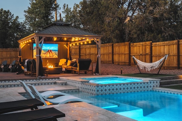 Un área de piscina con sillones reclinables, una zona de estar cubierta con una chimenea y una hamaca.