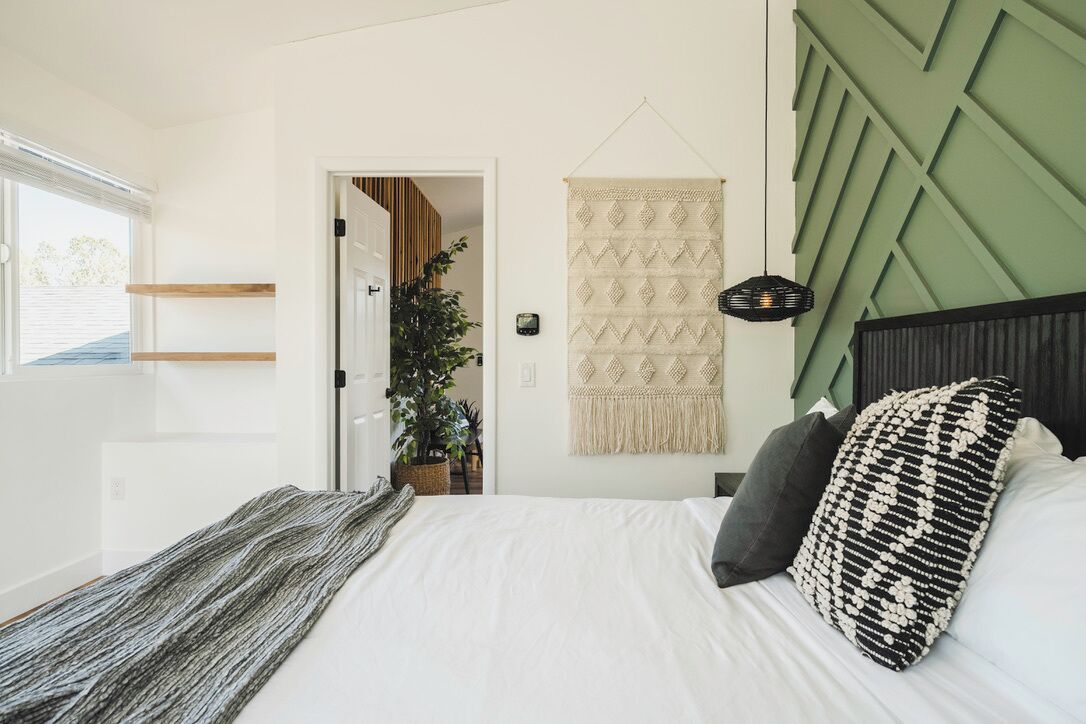 A bedroom with a bed, a green accent wall, a hanging macrame wall hanging, and a plant.