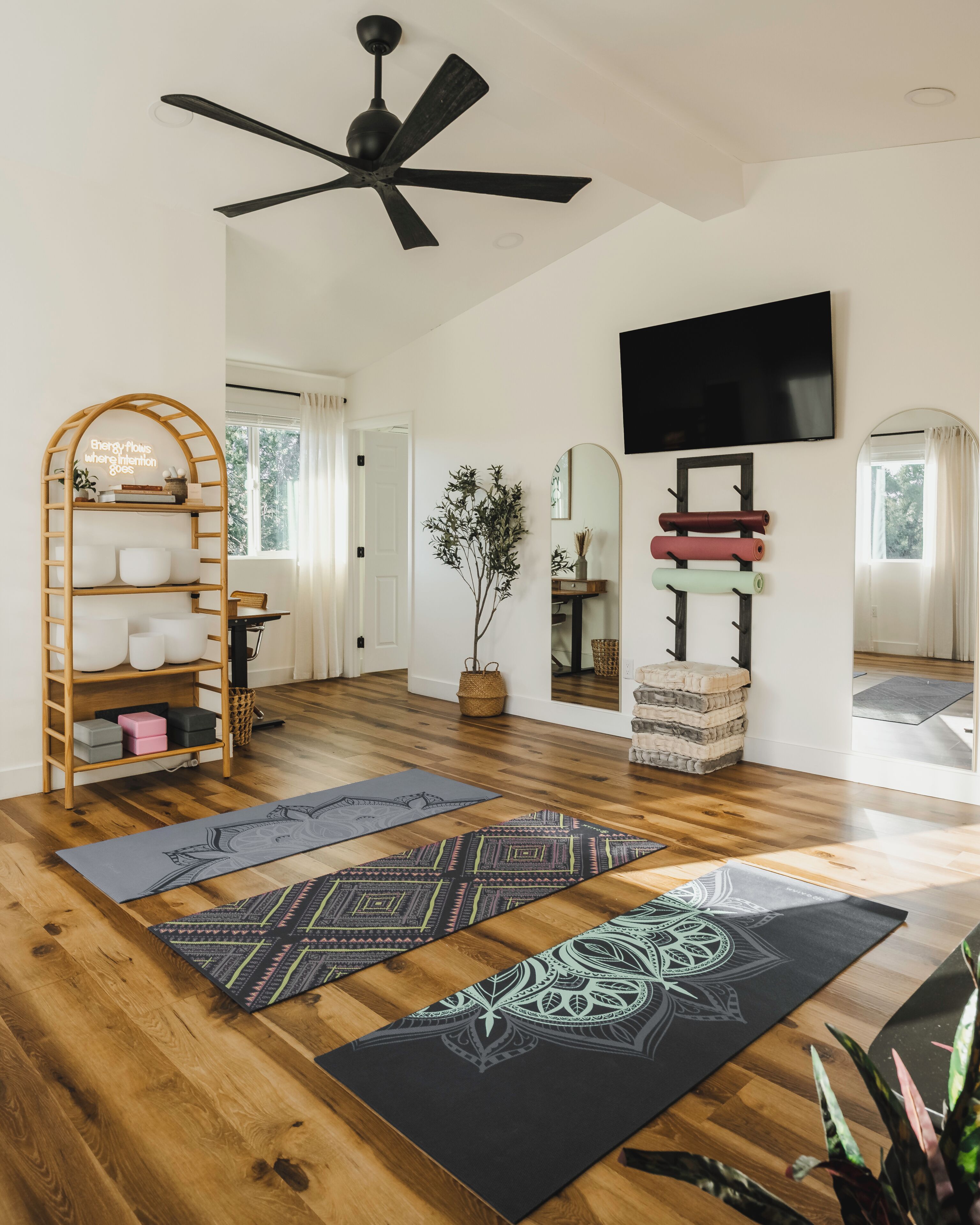 A spacious living room with a wooden floor, a ceiling fan, a TV mounted on the wall, and yoga mats on the floor.