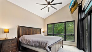 Un dormitorio con una cama grande, un cabecero de madera, una mesita de noche con cajones, un ventilador de techo y un ventanal con vistas a árboles.