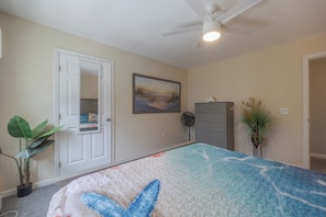 A bedroom with a bed, a door, a fan, a dresser, and a plant.