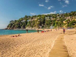 Uma praia arenosa com um calçadão de madeira, pessoas tomando sol e casas numa encosta ao fundo.