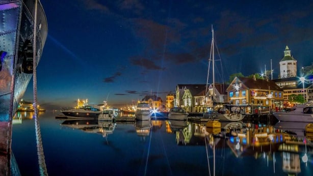 Un port de plaisance au crépuscule, avec des bateaux amarrés, des bâtiments le long du quai et une tour imposante en arrière-plan.