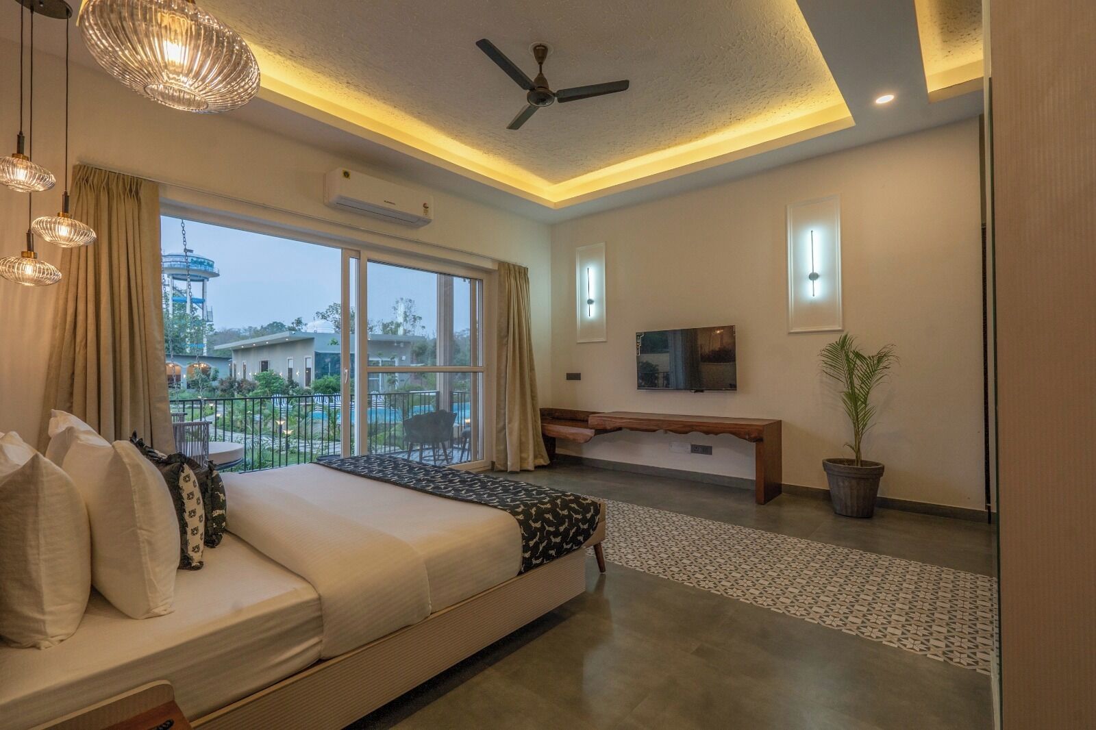 A modern hotel room with a large bed, a bench, a TV, and a balcony with a view of the pool and greenery.